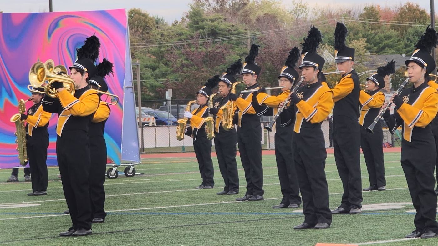 Woodwind and brass players performing on the field in front of a colorful backdrop