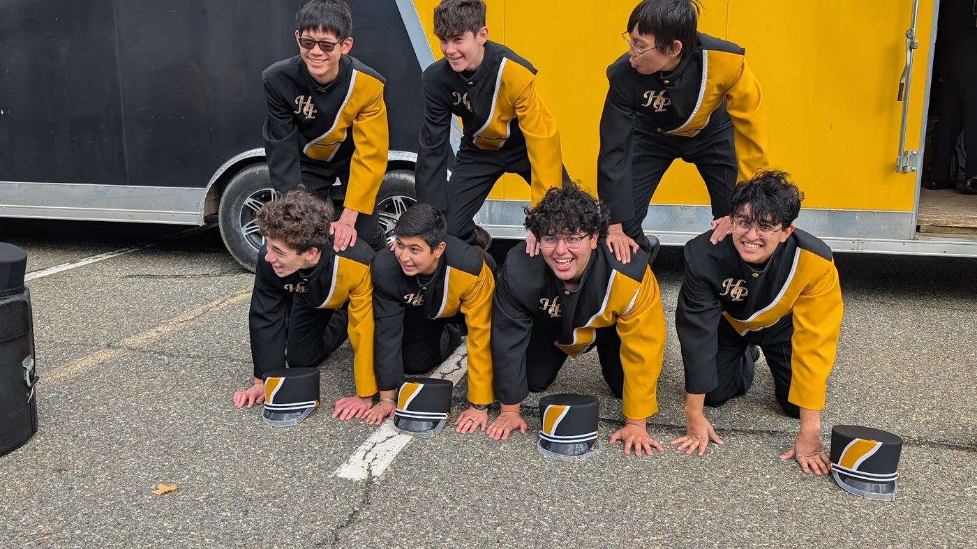 Seven band members laughing in a playful human pyramid with their hats on the ground